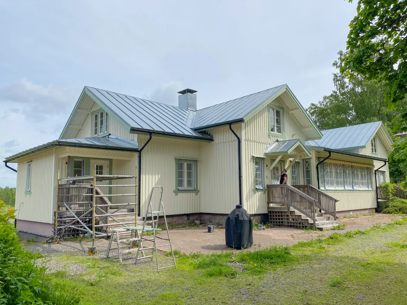 This image features a yellow wooden house with a person on a ladder working on the roof, set against a backdrop of greenery.