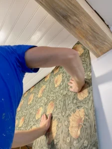 Hands of a person carefully applying floral patterned wallpaper to a wall.