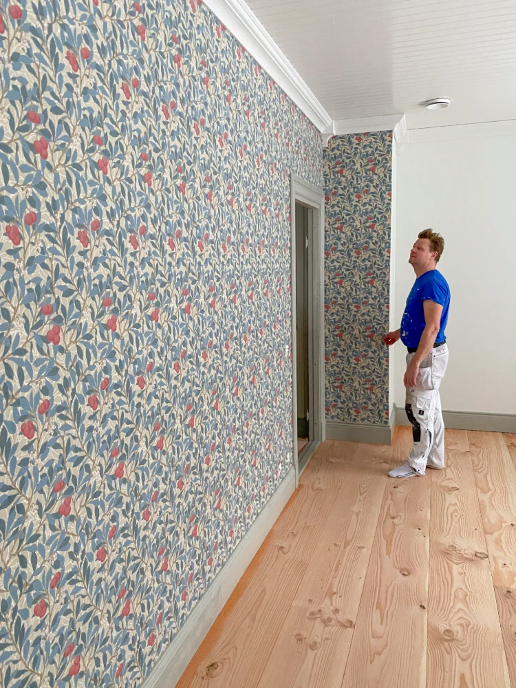 A person in a blue shirt and white pants stands in a room with patterned wallpaper featuring a floral design in shades of blue, red, and cream. The room has a wooden floor and appears to be undergoing some kind of renovation or decorating work.