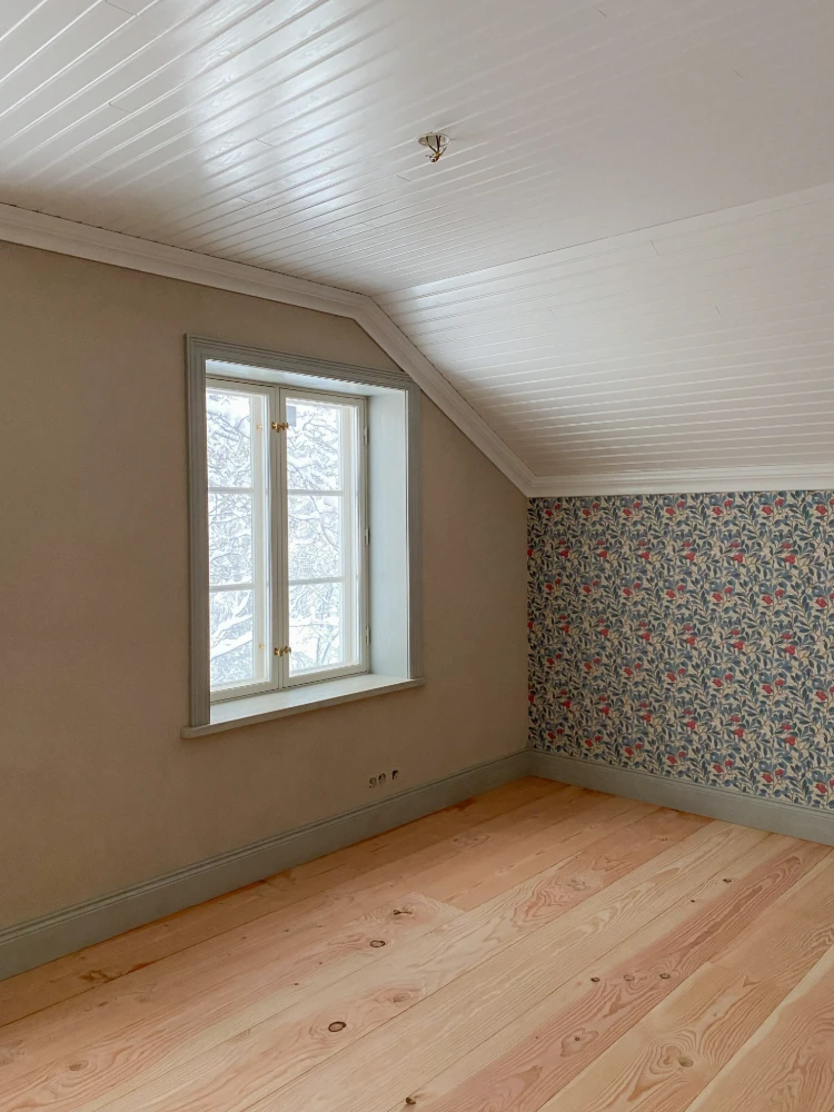 Interior room with wooden flooring, patterned wallpaper, and a window overlooking a snowy outdoor scene.