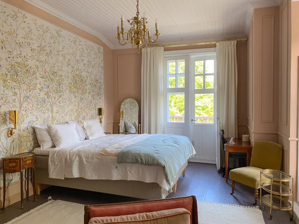 This image shows a cozy and charming bedroom with a floral wallpaper accent wall, a large bed with white linens, a chandelier overhead, and a window overlooking a scenic outdoor view.