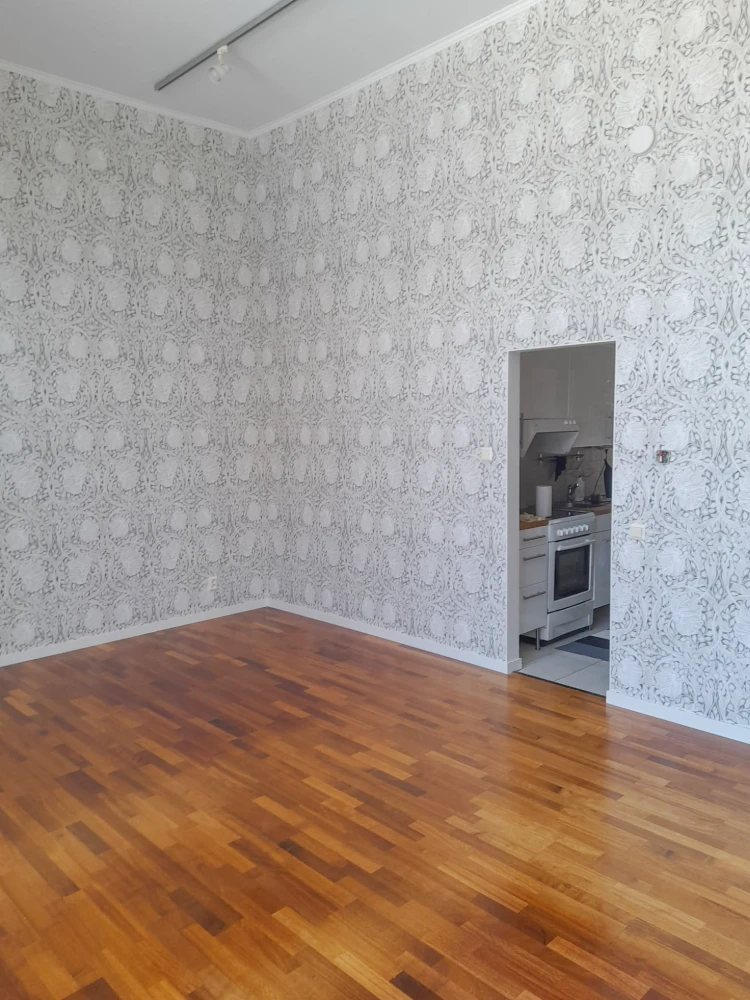 Empty room with patterned wallpaper and hardwood flooring, featuring a kitchen appliance visible through an open doorway.
