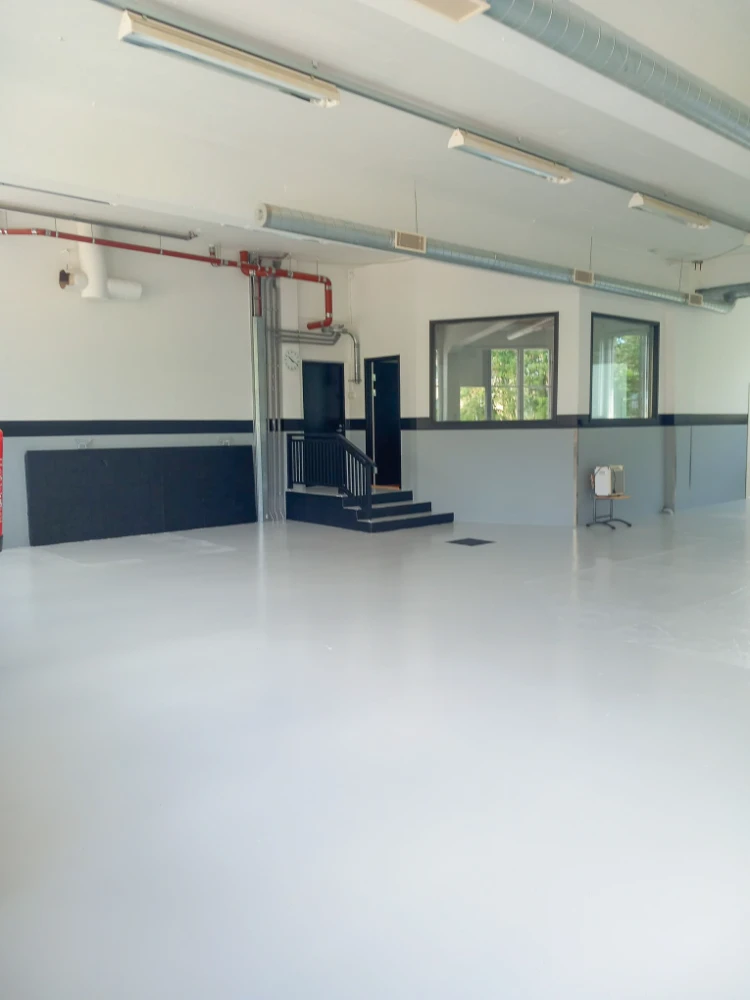 A large empty room with a white floor is depicted in the image. In the room, there is a man in a suit and tie standing near a lamp. The room has a white ceiling,