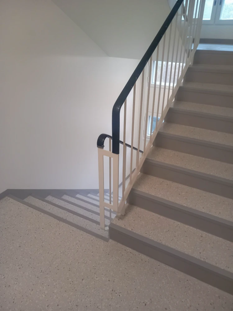 A staircase with a railing and a handrail is depicted in the image. The staircase is located in a room with white walls, and the railing is black in color.